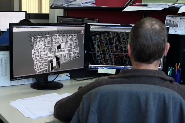 A man sits at a desk in an office, focused on two monitors displaying detailed architectural blueprints. Papers and office supplies are visible nearby.